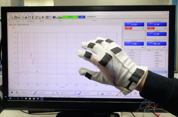 ETRI연구진이 그래핀 기반 직물형 스트레인-압력복합센서를 면장갑에 적용하여 손동작에 따른 신체신호 감지 기술을  인장 및 압축 측정 장비를 통해 시연하고 있는 모습 .